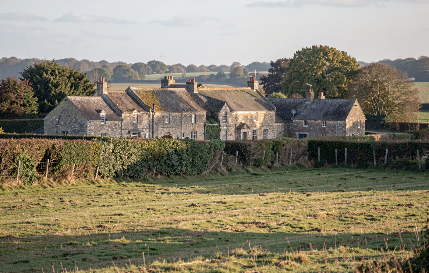 A stone hamlet in the Normandy bocage at low morning light, hedgerows and a mown meadow in the foreground, wide view from a slight rise.